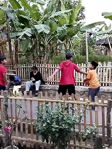 Five children are playing in a yard. A fence made of bamboo runs across the foreground. Behind the fence, two boys and three girls are gathered. One boy in a red shirt stands on the left, looking up and to the right. A boy in a blue shirt sits on a low wall in the center, looking down at a phone. A girl in a black shirt sits next to him, also looking at a phone. A girl in a red hoodie stands facing them. A boy in an orange shirt and a black cap stands on the far right, also facing them. A pink ball is thrown from the right towards the center, and the boy in the red shirt is attempting to catch it. Banana trees and other vegetation are visible in the background.