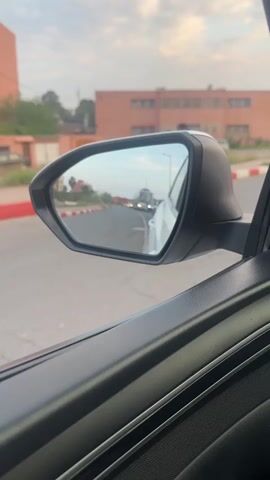 The video shows a close-up view from inside a car, looking out at the side mirror on the driver's side. The car appears to be stationary or moving very slowly on a road in Marrakesh, Morocco.

In the foreground, the black plastic of the car's interior door panel and the exterior side mirror casing are visible. The mirror itself reflects the road behind the car, showing a light-colored vehicle (possibly a truck or bus) followed by another car in the distance. The sky, which is partly cloudy and suggests either late afternoon or early evening, is also reflected in the mirror.

Outside the car window, to the left of the mirror, a person on a motorcycle passes by, moving from right to left. The road has distinctive red and white painted kerbs. In the background, there are buildings with a reddish-brown hue, characteristic of architecture found in Marrakesh. The overall scene depicts a typical street view from a car in an urban or semi-urban setting. There is no visible text.