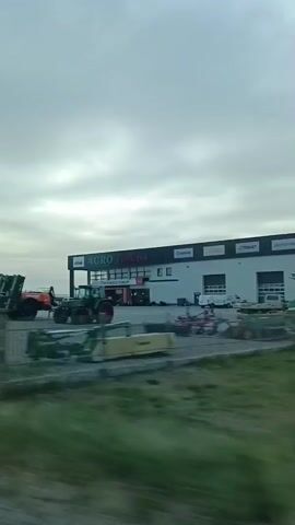 The video displays an outdoor view of an agricultural equipment dealership. In the foreground, agricultural machinery such as a red tractor with a yellow and red implement attached, a blue and red implement, and other various farm equipment are visible. Behind these, a green truck with a large green trailer is parked. A building with the sign "AGRO" is in the background, with several more pieces of farm equipment displayed in front of it, including a red water tanker. The sky is overcast with clouds. A fence runs along the bottom of the frame. In the distance, a wind turbine is visible in a grassy field. The footage appears to be shot from a moving vehicle, indicated by motion blur.