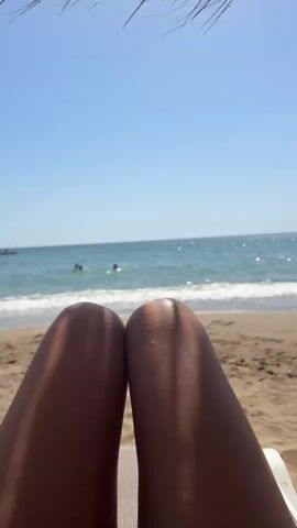 The video shows a first-person view of someone's legs resting on a beach chair at a beach. The camera is pointed upwards, with the legs filling the bottom half of the frame. Behind the legs, the ocean is visible with gentle waves rolling onto a sandy shore. The sky is clear and blue. In the distance, a few people can be seen in the water. The top of a straw umbrella or sunshade is visible at the very top of the frame, casting a shadow on the person's legs. The overall impression is one of relaxation and enjoying a sunny day at the beach.
