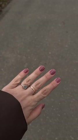 A woman's left hand is displayed against a gray, textured ground. The hand wears a brown jacket sleeve, a gold wedding band, and a gold ring with a blue stone. The fingernails are painted a mauve color. The hand is first shown with the fingers extended, then is clenched into a fist with the thumb extended upward.