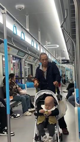 Inside a subway car, an elderly man in a navy and white patterned shirt pushes a stroller with a baby in it. The baby, who is bald, is holding something in their hands and looking down. The man is looking off to his right. Several other people are sitting on the blue seats lining the side of the subway car, some looking at their phones. The car is brightly lit by overhead lights, and there are silver poles with blue grab handles.  A white tote bag hangs from one of the stroller’s handles.