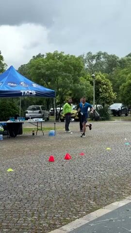 A man in a blue, short-sleeved athletic outfit is jogging along a cobblestone ground outside. He dodges around red and yellow traffic cones, and runs past a red yoga mat. In the background, there is a blue tent, cars parked in a grassy area, and a few men standing. The sky is overcast. The man is wearing a tag around his neck.
