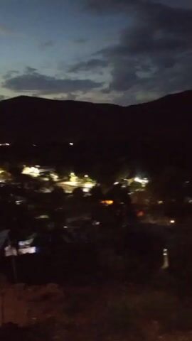 The video shows a dimly lit landscape at night. In the foreground, there is a makeshift shelter, possibly a tent or a small hut, illuminated by a warm light source inside. Beyond the shelter, a town or settlement is visible, with several buildings and streetlights dotting the landscape. The background is dominated by a silhouette of dark mountains against a twilight sky. Thin clouds are present in the sky, adding texture to the scene. The overall ambiance is quiet and peaceful.