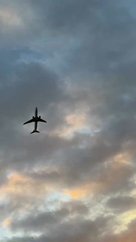 The video begins with a view of an airplane silhouetted against a cloudy sky. The clouds are various shades of gray with some patches of light orange. The plane moves out of the frame. The camera then pans down to show a metal fence with barbed wire at the top. Behind the fence is a grassy field with a bright orange sunset in the distance. The sun is partially obscured by the horizon.