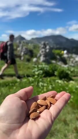 A close-up shot of a person's open hand holding a handful of almonds is in the foreground. The background is a blurry, green, mountainous landscape under a blue sky with scattered clouds. Several people with backpacks are walking on a trail in the middle ground, with one person in focus, a woman with a red backpack wearing a yellow shirt and shorts.