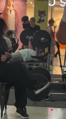 A video shows a person lying on a weight bench performing a bench press. Several people are standing around the weightlifter, some appearing to be spotters or spectators. The background suggests an indoor public space, possibly a gym or a store, with promotional displays visible, including a cartoon cat image. The person is lifting a barbell with considerable weight plates attached to each end. The lighting is bright, and the camera appears to be handheld, capturing the scene from a slightly elevated angle.