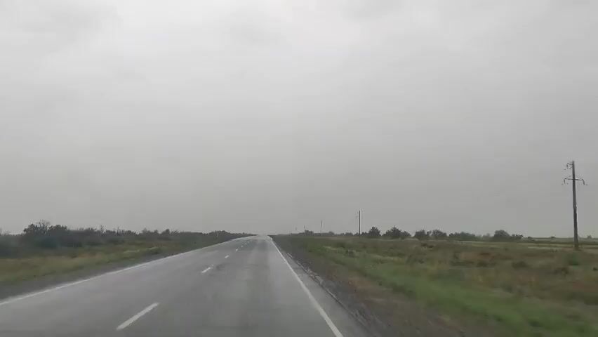 The video captures a point-of-view shot from a vehicle traveling on a wet asphalt road under a heavily overcast sky. The road stretches into the distance, with white dashed lines marking the lanes. To the left and right of the road are grassy fields with scattered bushes and some trees. Utility poles are visible in the distance on the right side of the road. The overall atmosphere is somber due to the gray, cloudy weather, suggesting it may be raining or has recently rained. The video appears to be a continuous shot of the road ahead, with no visible people or other vehicles interacting within the frame.