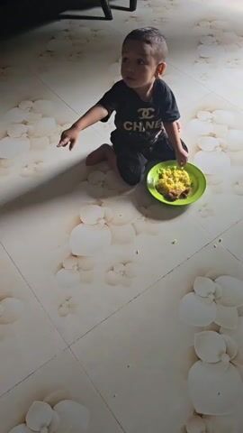 A young boy sits on the floor in front of a plate of yellow food. He is wearing a black t-shirt and black pants. He points to something off-camera. A cat walks across the floor in front of him. He then brings his hand to his mouth, as if eating. The floor is tiled with a floral pattern.