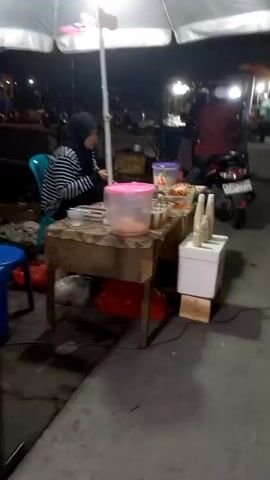 A nighttime street scene shows a food stall with a woman in a hijab and striped shirt serving food. A scooter with two passengers drives past, the rider wearing a red shirt and white cap, and the passenger a pink hijab. The stall has a table with various food containers and drinks. In the background, other food stalls and people are visible.
