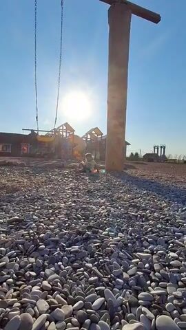 This video captures a sunny day at a playground in Timmolovo, Russia. The primary subject is a young child, dressed in a light green shirt and dark pants, lying on the ground amidst a wide expanse of smooth, grey pebbles. The child appears to be playing or resting near a large, rough-hewn wooden pillar that supports a swing set. The swing set's chains are visible, hanging from a wooden beam atop the pillar.

In the background, a playground structure with yellow and orange slides and climbing frames is visible. The sun is shining brightly in a clear blue sky, casting a warm glow and creating lens flare. The overall atmosphere seems peaceful and idyllic, suggesting a pleasant outdoor activity. The ground is covered entirely in pebbles, which are the most prominent textural element of the foreground. The presence of a playground, combined with the child's activity, points to a recreational setting. The time of day is clearly daytime due to the bright sun and clear sky. There is no visible text in the video.