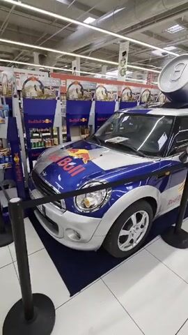 The video shows a Red Bull promotion in what appears to be a supermarket. A Red Bull-branded Mini Cooper car is displayed on a blue mat surrounded by black poles with silver tops. The car is primarily silver, blue and red, with the Red Bull logo prominently displayed. On top of the vehicle is an oversized model of a Red Bull can. Several Red Bull promotional display racks are behind the car. The camera circles around the car providing a view from different angles. The supermarket environment is visible in the background with various food and product shelves under bright overhead lights. There are no people visible in the video.
