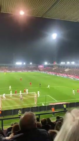 The video shows a live soccer match at night, viewed from the stands. The field is well-lit with artificial lights. Players in red and white jerseys are actively engaged in the game. A goal post is visible at one end of the field. Spectators fill the stands.