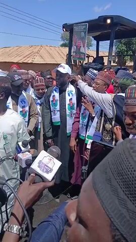 The video opens with a close-up of hands holding microphones, some with logos, indicating a media presence. The camera then pans up to reveal a group of men, most wearing traditional Islamic caps and some with sashes depicting faces. They are gathered outdoors, with a bright sky and buildings visible in the background. One man in the center, wearing a white baseball cap and a sash, is speaking into a microphone. Another man to his right, in a maroon outfit and a similar sash, appears to be responding or interacting with the crowd. Behind them, a large piece of machinery, possibly a tractor, is visible with a banner attached to it, also featuring faces and text. The overall atmosphere suggests a public event, likely a political rally or press conference, given the presence of media and campaign-like attire. The focus is on the men speaking and the media documenting the event.