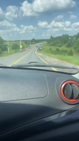 The video is a first-person perspective from inside a vehicle traveling on a road. The camera is pointed forward, capturing the view through the windshield. The dashboard of the vehicle is visible in the foreground, with a dark textured surface and a red-rimmed air vent on the right. Outside, a paved road with yellow lane markings stretches ahead, curving gently to the right. The road is lined with a barrier made of alternating red and white plastic elements on the left and a metal guardrail on the right. Beyond the road, rolling green hills and trees are visible under a bright blue sky dotted with fluffy white clouds. In the distance, a few cars can be seen on the road. A blue road sign with white lettering and a speed limit sign indicating 20 are visible on the right side of the road. The overall scene suggests a sunny day in a rural or suburban area.