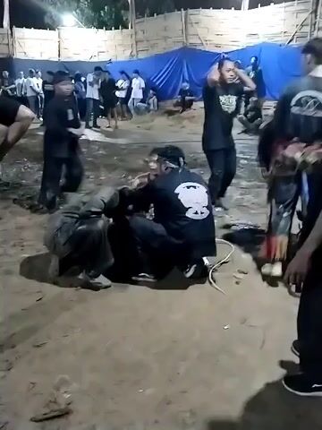 The video captures an outdoor nighttime scene in Jenggrik, Indonesia, featuring a group of people gathered for what appears to be a cultural event, likely related to Reog Ponorogo. The setting is an open area with bare earth, which looks somewhat muddy. Artificial lighting illuminates the scene.

In the foreground, two individuals are seated on the ground. One person, wearing a black shirt and pants, appears to be in a state of distress, looking to the side. Another person, also in a black shirt with a white circular emblem on the back, is sitting closely, attending to or comforting them. A white rope or cable is visible on the ground nearby.

Surrounding these central figures are several other people, predominantly males, mostly dressed in black t-shirts and pants. One person in the immediate foreground, facing away from the camera, wears a black t-shirt with a detailed gold design on the back that reads "PAGUYUBAN SENI REOG TURONGGO JATI," indicating an affiliation with the traditional Indonesian Reog Ponorogo performance art. Another person in a black t-shirt and pants stands to the right, observing. On the far right, part of a traditional horse costume, characteristic of Reog Ponorogo, is visible.

In the background, more people are gathered, standing or sitting. Behind them, a large blue tarpaulin serves as a backdrop or temporary wall, with wooden or bamboo fence-like structures further behind. The event appears to be a communal gathering, possibly involving trance or intense emotional states often associated with such cultural performances.