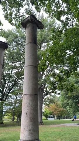 The video pans across a row of large, weathered stone columns situated on a grassy lawn. The columns appear to be ancient Greek or Roman in style, with fluted shafts and ornate capitals. They are surrounded by lush green trees, and the sky is overcast. In the distance, a modern building can be seen, and a car is parked nearby. The overall impression is one of a serene and historic setting, with a sense of timelessness conveyed by the imposing columns. There are no people visible in the video, and no actions or interactions are depicted. The video is static, with no camera movement or subject motion.