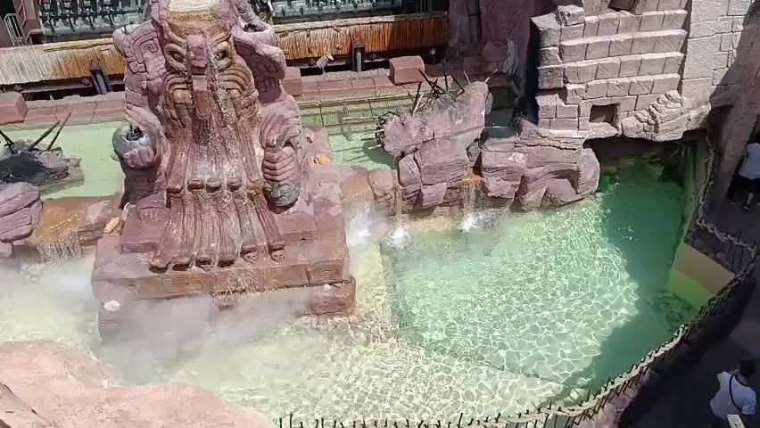The video captures an aerial view of an amusement park ride with a Mayan-themed aesthetic. A large, reddish-brown stone structure resembling a deity's face with elaborate carvings is the centerpiece. Water cascades down from various points around the structure into a pool with clear turquoise water. Steam or mist emanates from the base of the deity statue. In the background, a roller coaster train with passengers seated in rows is visible, moving along a track. Rocky formations and stone walls, also with carved patterns, surround the pool area. Several people are seen on the ride and in the background, though their specific actions are not clear from this elevated perspective. The overall environment suggests a hot, sunny day.