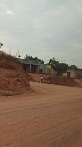 The video captures a scene of a dirt road with buildings and people. The road is composed of reddish-brown dirt. On the side of the road, there are buildings, trees, and banana plants. Two men are walking along the side of the road. The sky is overcast with a grey tint. The camera appears to be mounted inside a moving vehicle.