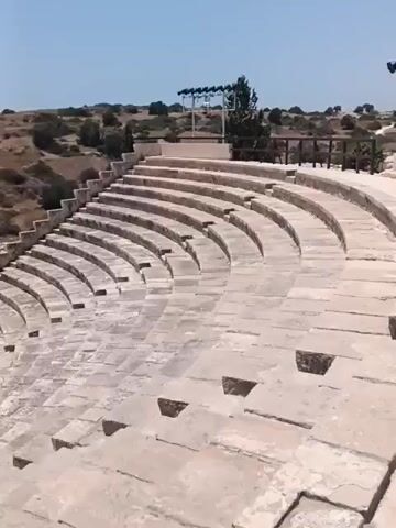 The video shows an outdoor amphitheater with tiered stone seating. In the background, there is a hillside covered in dry brush and a clear blue sky. To the left of the amphitheater is a rectangular pool with a dark surface, bordered by a wooden fence. A person is walking in front of the pool. Beyond the pool and hillside, the turquoise ocean is visible.