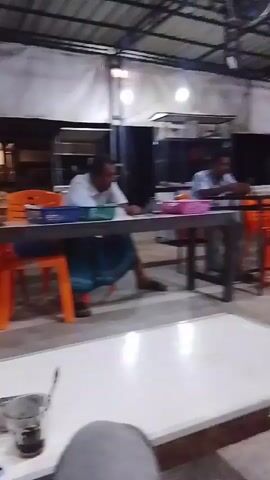The video shows an indoor scene, likely a restaurant or community hall with an open-air section. Two men are seated at tables. The man on the left wears a white shirt and a sarong, and appears to be looking down. The man on the right wears a light-colored shirt and pants and is also looking downwards. Another man, wearing a white cap, white shirt, and plaid sarong, is seated at a table in the foreground, looking down and resting his hands on the table. There are several plastic chairs of orange and red colors around the tables. The environment appears to be dimly lit, with overhead lighting and some natural light from the outside. In the background, there are shelves, water dispensers, and what appears to be a motorbike parked outside. The camera is handheld and the footage is somewhat blurry.