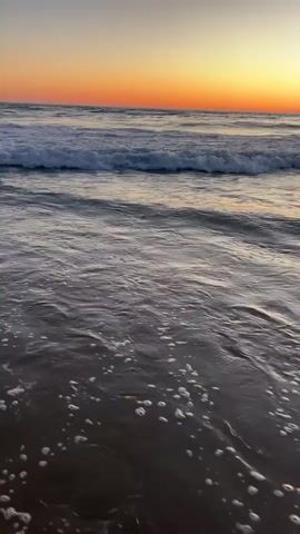 The video shows a close-up of ocean waves crashing on a beach at sunset. The sky is a gradient of orange and purple, with the sun low on the horizon. The water is dark and reflects the sky's colors. Foamy white waves roll towards the shore, with smaller waves and ripples visible closer to the camera. The sand is dark and wet. The video captures the movement and sound of the waves.