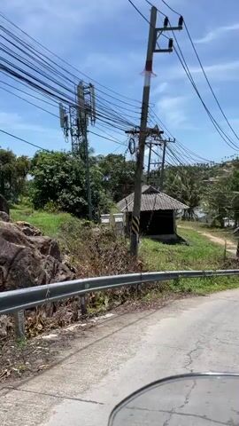 The video shows a view from a moving vehicle driving along a road. The foreground shows a gray metal guardrail. Beyond the guardrail is a grassy area with a small, derelict shack with a gray tile roof.  To the left is a cell phone tower. Power lines crisscross the sky above. Palm trees and other vegetation are visible further in the background. The sky is blue with some clouds. A glimpse of the ocean can be seen in the distance between the trees.