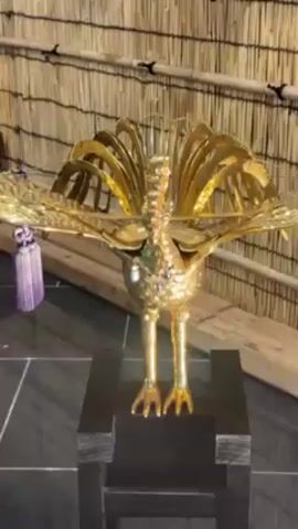 A golden phoenix statue stands on a black pedestal against a bamboo-like wall with decorative items. The statue has its wings spread wide and is adorned with intricate details. The scene appears to be indoors, possibly in a cultural or display setting. There are sounds of people talking in the background.
