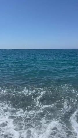 The video shows a view of the ocean from a pebble beach. The sky is clear and blue. The water is a vibrant turquoise color, with small waves gently lapping at the shore. The pebbles on the beach are gray and varied in size. There are a few distant specks that might be boats or buoys on the horizon. The overall impression is a calm and serene coastal scene on a sunny day.