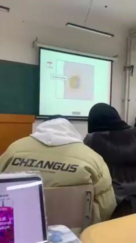 A classroom scene shows a projection screen displaying a diagram of a cell.  Several students are seated in the classroom, including one wearing a tan jacket with the word "CHIANGUS" printed on the back. A laptop is visible in the foreground with what appears to be a zoomed-in image of a cell. The teacher is giving a lecture about the cell. A student in a black hijab is sitting in front of the camera to the right. The overall scene suggests an educational setting during a lecture.