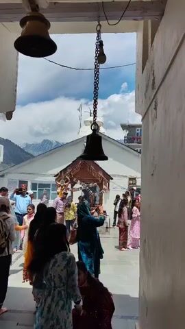 This is a vertical, medium shot video, taken from a doorway, of a group of people gathered outside a temple structure. Three bells are visible, two hanging from the doorway ceiling and one from a chain in the center of the frame. In the background, there is a white temple-like building with a brown wooden archway in front of it. Several people are walking in and around the temple area. The sky is cloudy with visible mountains in the distance. The people are dressed in colorful clothing, and some appear to be taking pictures. One woman in the foreground, wearing a dark red outfit, is raising her arm and touching one of the bells.