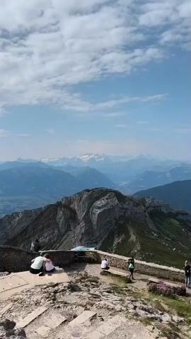 The video shows a panoramic view from a mountaintop, looking out over a vast mountain range under a clear blue sky with some scattered clouds. In the foreground, several people are visible on a paved area with stone walls. Two individuals are sitting on a bench, facing away from the camera, while another person is standing nearby. Further down the path, a couple of other people are walking. A steep, rocky mountain peak dominates the middle ground. Beyond this, layers of progressively distant mountains fade into a blue haze. Some of the furthest peaks are capped with snow and ice. The environment appears to be a popular scenic overlook.