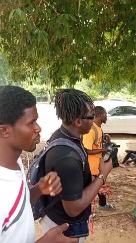 Here is a forensic description of the video:

**Scene:** The video appears to be taken outdoors, likely in a public space, with a dirt ground and trees overhead.

**People:** There are several individuals present, mostly men, dressed in casual clothing. Their attire varies, including t-shirts, traditional African garments (green robe), basketball jerseys (Bulls), and tracksuits. Some wear hats, sunglasses, and backpacks. Several of them appear to be using mobile phones.

**Objects:** The scene includes a few parked cars on the roadside and a parked motorcycle.

**Actions and Interactions:** The people are standing or sitting around a tree. Some of them appear to be interacting with each other in a casual manner, possibly in conversation.  A man is sitting on the motorcycle. One man is holding his phone and wearing a backpack, and a red string.

**Environment:** The environment is a mix of natural and urban elements with a road in the background. The presence of trees suggests a park or public area.

**Overall:** The video depicts a group of men casually gathering outdoors, possibly socializing or waiting for something, with the environment suggesting a public space or park.