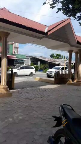This short video captures a street scene in Pulokiton, Indonesia, during daylight hours under a cloudy sky. The foreground features the rear end of a parked motorcycle, its black seat and some yellow accents visible. Beyond it, a paved area leads to a covered walkway supported by decorative pillars.

In the middle ground, a dark-colored SUV is parked on the right side of a road. Across the road, a white car is visible, suggesting traffic flow. Buildings with red roofs and some greenery line the street. The architecture suggests a tropical setting.

The overall atmosphere appears calm and ordinary, with no distinct events or emotional cues. The presence of parked vehicles and buildings indicates a commercial or residential area. There is no discernible text visible in the video.