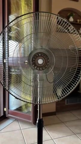 The video shows a close-up, static shot of a standing fan. The fan has a metal grill and blades, and is mounted on a black stand. The background is blurry, but appears to be an indoor setting with a tiled floor and large glass doors that lead outside. Sunlight is streaming through the doors and reflecting off the fan's grill. There are some shelves visible in the background with decorative items, and a clock on the wall. The fan's blades are in motion, indicating it is turned on. The scene appears to be daytime, and the lighting suggests it might be a warm day. No people are visible in the video. There is no visible text in the video.
