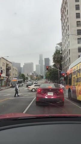 This media file captures a street scene in Los Angeles, USA, likely during the daytime under an overcast sky. The primary focus is a traffic intersection, with several vehicles stopped at a red light.

In the foreground, we see the interior of a red car, presumably the perspective of the person recording. A dark red Toyota Corolla with California license plate "9JSH056" is directly ahead, also stopped. To the right, a bright yellow and orange bus with advertisements is partially visible.

Across the intersection, several other cars are stopped, including a white car on the far left and a white SUV further down the road. A pedestrian, dressed in dark clothing and holding a cane, is walking across the crosswalk, seemingly unimpeded by the traffic. Another pedestrian can be seen on the sidewalk to the right.

In the background, the distinct Los Angeles skyline is visible, dominated by tall skyscrapers, including the U.S. Bank Tower. Palm trees line the street, adding to the urban landscape. Street signs are visible, including one that reads "WITMER." The traffic lights are red, indicating a stop. The overall atmosphere appears to be that of a typical city commute, with the cloudy weather lending a slightly subdued tone to the scene. The presence of multiple vehicles and pedestrians suggests a moderately busy urban environment.