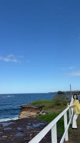 The video captures a panoramic view of a coastal landscape under a clear blue sky with a few scattered clouds. The foreground features a rocky shoreline with large, dark, wet rocks, suggesting recent wave activity. A white railing runs along the right side of the frame, indicating a walkway or viewing area. Beyond the rocks, the ocean stretches out, displaying various shades of blue with white-capped waves crashing against the shore. A slightly elevated cliff face with layered rock formations, covered in sparse green grass, is visible on the left side of the ocean. In the distance, a faint line of land or buildings is discernible along the horizon. The overall impression is one of a serene and natural coastal environment.