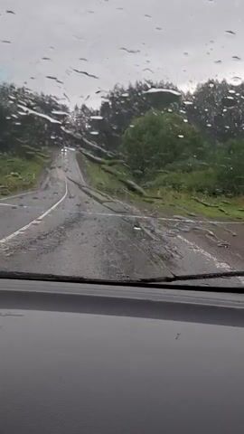 The video shows a view from inside a vehicle, looking through a rain-streaked windshield at a wet, paved road. The road is surrounded by green trees and vegetation. The sky is overcast. There appear to be some fallen branches or debris on the road. In the distance, a white line is visible across the road, potentially indicating a roadblock or caution. There are no people clearly visible in the video. The overall impression is a dreary, wet, and potentially hazardous driving condition.