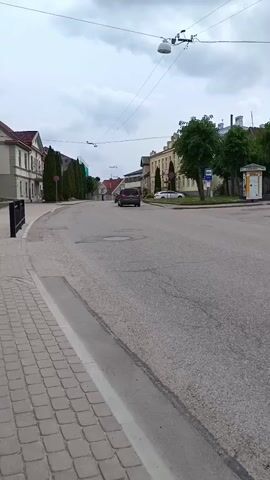 The video presents a wide, static shot of a street corner in the town of Aizpute, Latvia, under an overcast sky during daytime. The asphalt road curves gently to the left in the foreground. A dark sedan drives away from the camera at the beginning of the clip, moving into the distance.

On the left side of the frame, a cobblestone sidewalk is separated from the road by a curb. A person in light clothing walks away from the camera along this sidewalk between the 0:02 and 0:03 mark. Beyond the sidewalk, a row of tall, narrow evergreen trees lines the street, fronting multi-story buildings with traditional European architecture.

On the right side of the street, similar buildings are visible, interspersed with deciduous trees. A standard blue and white bus stop sign is clearly visible, positioned near a bus shelter that has posters or advertisements on its side, though the text is unreadable. A white car is parked further down the street on the right. Overhead wires and streetlights are strung across the street above. The scene depicts a quiet, typical street view of a European town with light pedestrian and vehicular activity.