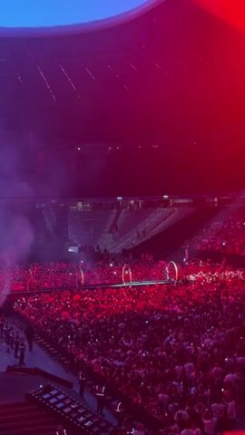 This is a vertical, high-angle, long-shot video of a large concert or event taking place in a stadium. The stadium is filled with a dense crowd of people, most of whom are holding up their phones. The lighting is dramatic, with a mix of red and blue hues. Smoke or fog is present, adding to the atmospheric effect. In the foreground, a stage area is visible with what appears to be audio equipment. Further back on the stage, a tall, metallic, humanoid figure is illuminated. Behind the figure, a large screen displays a profile image, possibly of a performer. Two circular, illuminated hoops are positioned on the stage, and it appears a performer may have just entered or exited through one of them. Security personnel in uniforms are visible at the edges of the crowd and near the stage. The upper portion of the stadium, including seating areas and the ceiling structure, is visible and also illuminated by the colored lights.