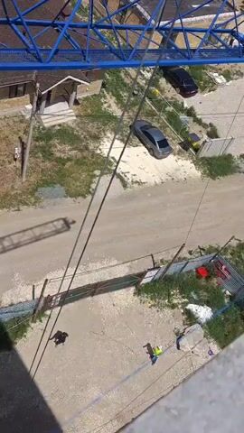 The video shows a construction site from a high vantage point, likely from a crane. The primary focus is on a large blue crane assembly. In the first few seconds, the view pans across a residential area with houses and then shifts to the construction site. The crane's boom is being assembled or dismantled, with sections being lifted and connected. There appear to be workers on the crane itself. The ground below shows a construction area with materials, vehicles, and fences. The environment is outdoors, with a blue sky and clouds visible.