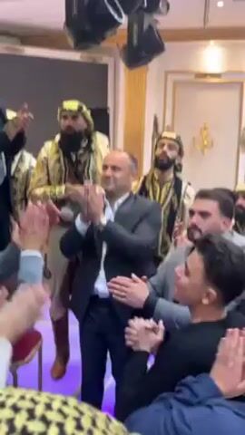The video depicts a group of men gathered in an indoor space, possibly a wedding or celebration venue. Several men are clapping their hands, indicating a celebratory atmosphere. Some men are dressed in formal suits while others are wearing traditional attire, including ornate robes and head coverings, suggesting a cultural performance or ritual. The environment is decorated with lighting fixtures and ornate architectural details. The overall impression is that of a joyous and culturally rich gathering.