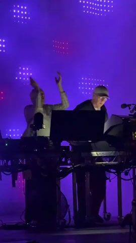 A short video clip shows two individuals on a stage during what appears to be a performance. The individual on the left, wearing a plaid shirt over a white t-shirt, is raising their arms in the air with eyes closed, seemingly reacting to the music. The individual on the right, wearing a black t-shirt and a baseball cap, is focused on a laptop and paperwork at a console. Behind them, a large LED screen displays a pattern of white and red lights. The stage is lit with blue and purple lights, and smoke or haze fills the air. The scene is dimly lit, with spotlights focused on the performers. The video transitions between a brightly lit blue stage and a darker, smokier environment.