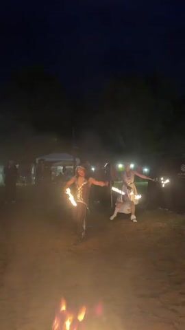Two people are fire dancing outdoors at night. One person in a dark outfit with a gold top is in the foreground, spinning fire around their body, occasionally breathing fire. The other person in a white outfit is in the background, also spinning fire with sticks. There's a small fire on the ground between them. In the background, other people are watching, and there are tents and trees. The overall setting is dark with light from the fire and distant lights.