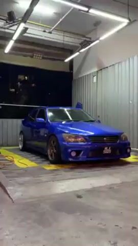 The video shows a blue Lexus IS200 parked in a car wash. The car has aftermarket wheels and body kit. The interior lights are on, and there is an open door on the passenger side. The car wash is well-lit with fluorescent lights. There is a hose with an orange cord.