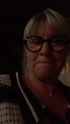 A woman with blonde hair and glasses is in the frame. She is wearing a black and white striped top. The lighting changes from dark to illuminated with colorful lights and then back to dark.