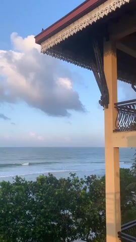 This video is a landscape shot of a tranquil ocean view at sunrise or sunset. The camera is positioned on a balcony or upper floor of a building, with part of the building's structure visible in the foreground, including a brown-tiled roof and a beige-colored pillar with ornate metal railings. Below the building, lush green trees and palm fronds are visible. The ocean stretches out to the horizon, with gentle waves rolling onto the shore. The sky is a clear blue with scattered, fluffy white clouds, some of which are tinged with the warm colors of the setting or rising sun. The overall mood is peaceful and serene. There are no people visible in the video.