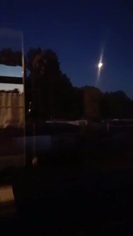 This media file captures a dark, nighttime scene, likely taken from inside a vehicle looking out through a window. The dominant subject is the deep blue night sky, with a single, bright streetlamp casting a glow upwards. Silhouetted against the sky are the dark shapes of trees and a building, suggesting a suburban or semi-rural setting. The foreground is very dark, obscuring most details, but reflections on the window pane are visible on the left side. These reflections suggest an interior with some kind of fabric blinds or curtains and possibly other objects.

The time of day is clearly night, as indicated by the dark sky and artificial lighting. There are no people or active events depicted. The overall mood is quiet and still. While no explicit text is visible, the presence of the streetlamp and building implies a developed area. Given the location context of Mahilyow, Belarus, this could be a view of a residential or commercial area outside the city center at dusk or after dark. The darkness and the single light source create a somewhat solitary or introspective atmosphere. The slight blurriness of the image might be due to movement of the camera or low light conditions.