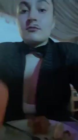 A person is sitting at a table in what appears to be a dining area, possibly a restaurant or a banquet hall. They are wearing a black blazer over a white top, and they also have a bow tie. A fake mustache is applied to their face.  On the table in front of them is a plate with food, including what seems to be a piece of fried food and a lemon wedge.  The person raises a champagne glass, takes a sip, and then looks directly at the camera. The background is somewhat blurred, but there are hints of other furniture and decorations, including a cabinet or side table with colorful inlays. The lighting is warm and somewhat dim. The music in the background is loud and upbeat.