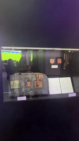 The video pans across a museum exhibit. In the foreground, a glass display case contains a vintage-looking suitcase adorned with stickers and patches. Behind the suitcase, also in the display case, are two crest-like objects. To the right of the suitcase, on a white stand, are two documents. Further to the right, a wall with text and images is visible, featuring the heading "THE WILDERNESS YEARS 1951-1986". A second display case contains a blue jacket and hat, likely part of the exhibit. The lighting in the exhibit is dim and atmospheric, with purple hues present. Several blurred figures of people can be seen reflected in the glass cases and in the background, suggesting the exhibit is open to the public.
